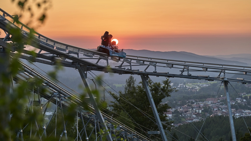 Cubea – privater Rückzugsort im Bayerischen Wald Zwei Personen auf einer Sommerrodelbahn bei Sonnenuntergang mit Bergblick